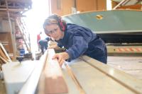 Eine Bootsbauerin prüft in ihrer Werkstatt ein geschnittenes Holzstück.